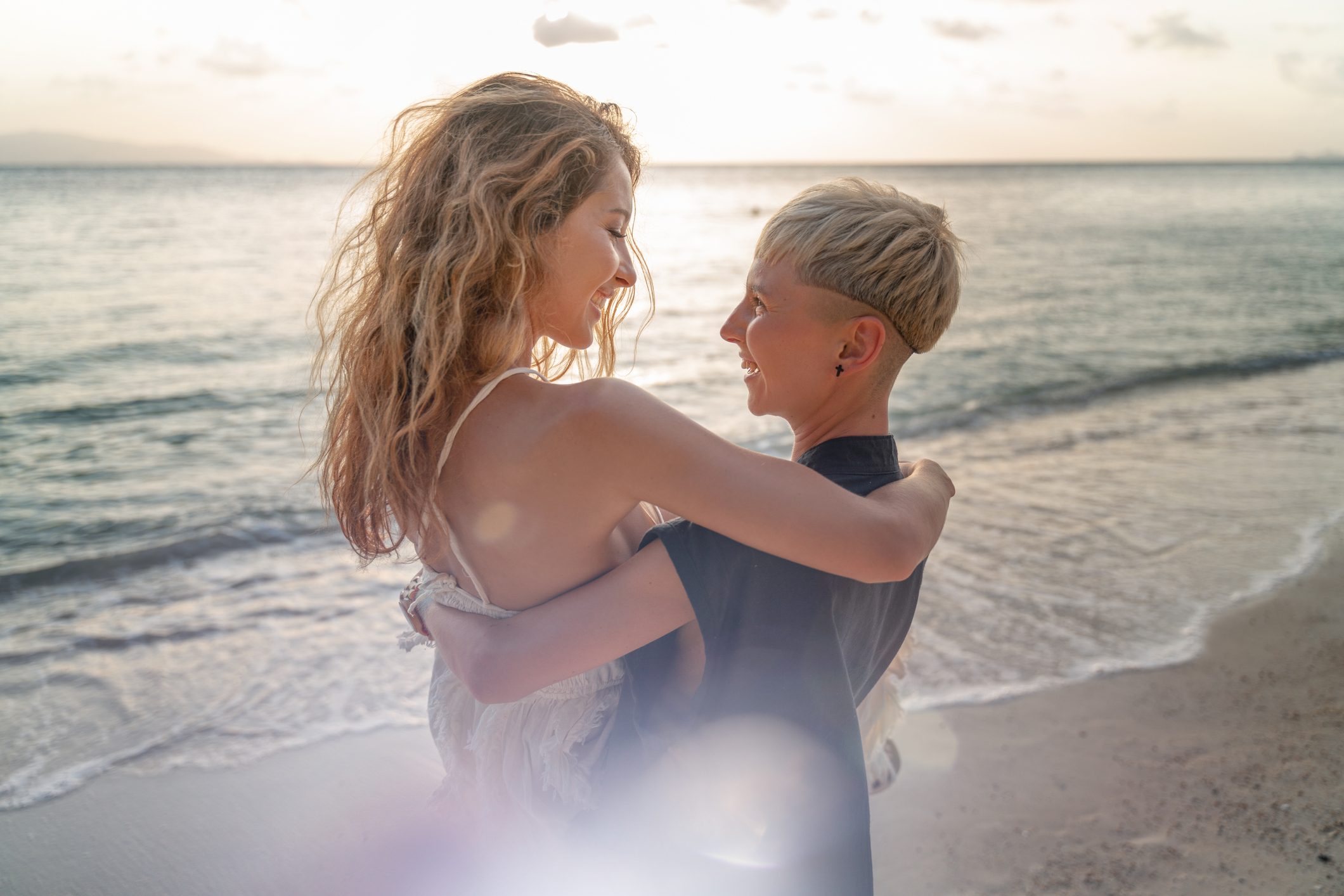Beautiful girl carries her wife girlfriend in her arms, during a wedding and a honeymoon on the seashore at sunset, lesbian wedding and LGBT community rights